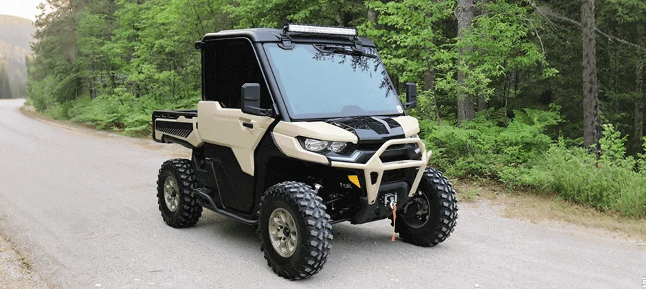 A tan Can-Am Defender HD10 UTV equipped with a winch and light bar parked on a wide dirt trail in a forest.