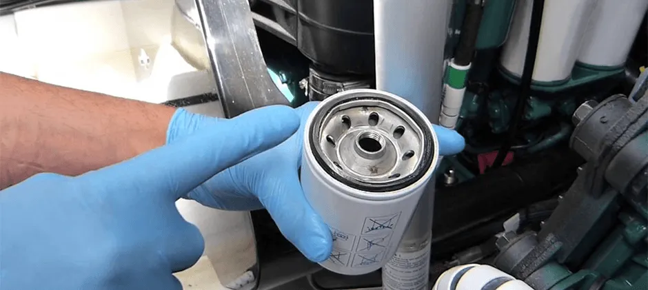 A person wearing gloves performs a DIY fuel filter replacement on a heavy equipment engine, holding a new aftermarket filter.