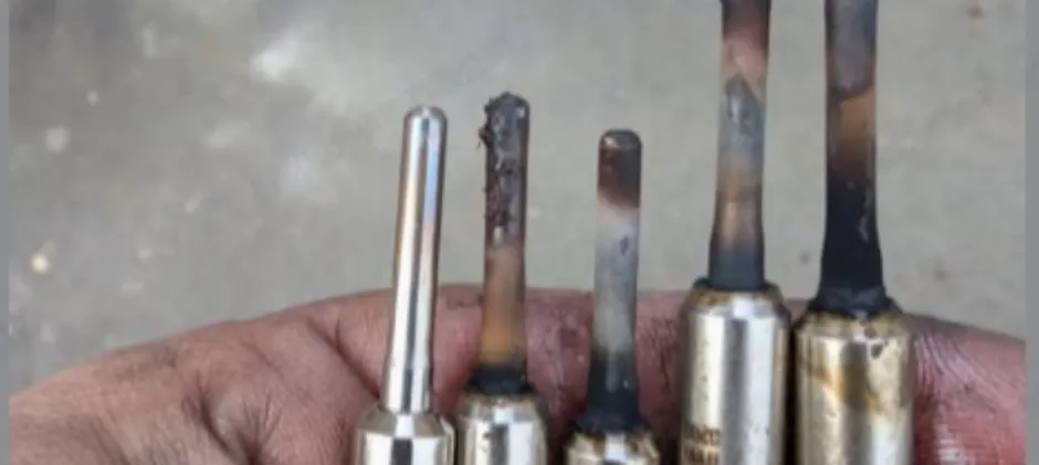 A mechanic's hand holding five glow plugs, showing a new plug next to four used ones with carbon buildup, a broken tip, and heat discoloration.